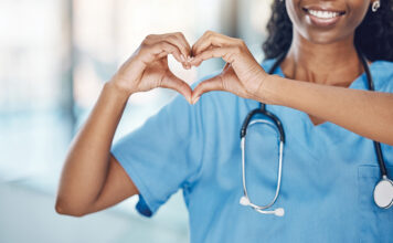 Closeup african american woman nurse making a heart shape with her hands while smiling and standing in hospital. Take care of your heart and love your body. Health and safety in the field of medicine