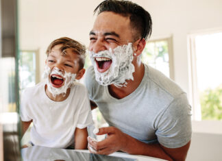 Man and little boy with shaving foam on their faces looking into the bathroom mirror and laughing. Father and son having fun while shaving in bathroom.