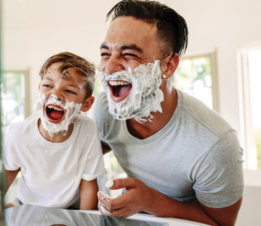 Man and little boy with shaving foam on their faces looking into the bathroom mirror and laughing. Father and son having fun while shaving in bathroom.