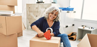 Middle age grey-haired woman smiling happy packing kitchen cardboard box at new home.