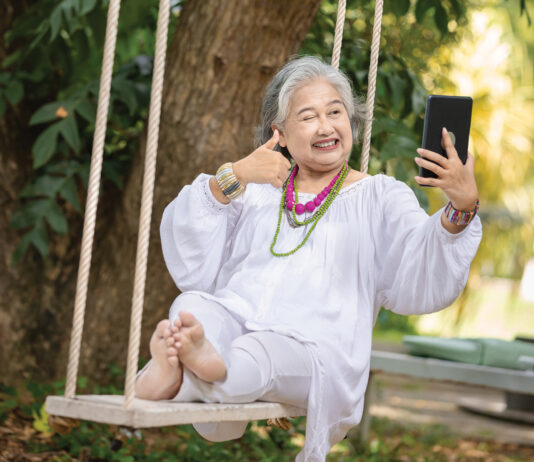 Elderly Woman Relaxing on a Swing While Using a Tablet in a Serene Outdoor Setting, Surrounded by Lush Greenery and Natural Beauty