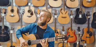 Young man plays on acoustic guitar in music store. Assortment in musical instruments shop, male musician buying equipment