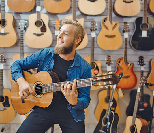 Young man plays on acoustic guitar in music store. Assortment in musical instruments shop, male musician buying equipment