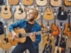 Young man plays on acoustic guitar in music store. Assortment in musical instruments shop, male musician buying equipment