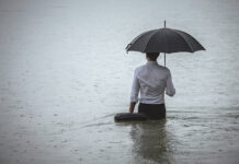 Rear view on a handsome man wearing white shirt and holding umbrella and a briefcase, standing in the water on a rainy day. The man waded knee-deep in water. Selective focus, three quarter length, copy space has been left.
