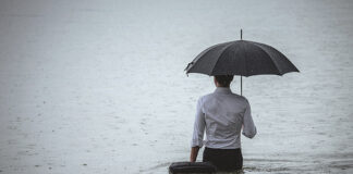 Rear view on a handsome man wearing white shirt and holding umbrella and a briefcase, standing in the water on a rainy day. The man waded knee-deep in water. Selective focus, three quarter length, copy space has been left.