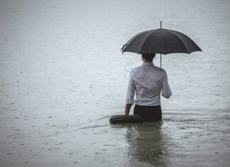 Rear view on a handsome man wearing white shirt and holding umbrella and a briefcase, standing in the water on a rainy day. The man waded knee-deep in water. Selective focus, three quarter length, copy space has been left.