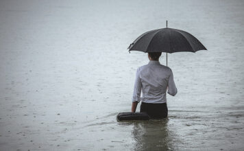 Rear view on a handsome man wearing white shirt and holding umbrella and a briefcase, standing in the water on a rainy day. The man waded knee-deep in water. Selective focus, three quarter length, copy space has been left.