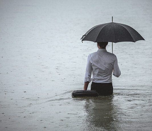 Rear view on a handsome man wearing white shirt and holding umbrella and a briefcase, standing in the water on a rainy day. The man waded knee-deep in water. Selective focus, three quarter length, copy space has been left.