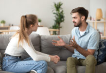 Serious young couple sitting together on sofa, talking about relationships, spending time together at home, focused wife listening to speaking husband, friends having conversation