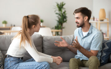 Serious young couple sitting together on sofa, talking about relationships, spending time together at home, focused wife listening to speaking husband, friends having conversation